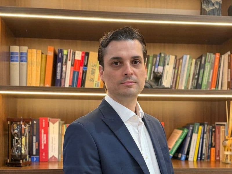 O advogado, de terno azul-marinho, em pé diante da estante de livros jurídicos do escritório.